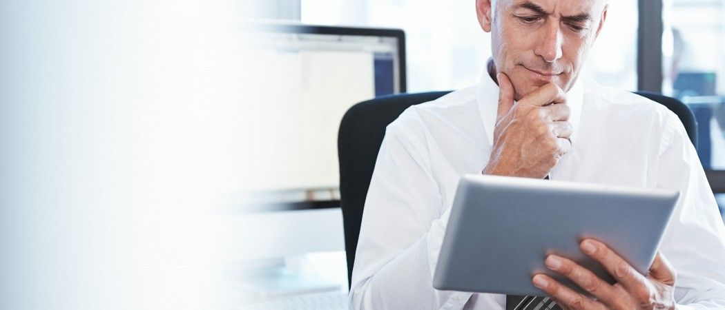 Ein Mann in einem Anzug schaut auf ein Tablet und denkt nach. Er sitzt an einem Schreibtisch mit einem Computermonitor.