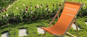 Ein Garten mit grünem Rasen und einer Reihe roter und weißer Tulpen. Ein orangefarbener Liegestuhl mit #meinWeinviertel steht auf dem Rasen. Im Hintergrund befindet sich ein Holzzaun mit einem Teich.