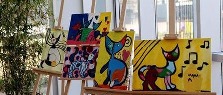 Mehrere Gemälde von Katzen sind auf Staffeleien vor einem Glasfenster ausgestellt. Eine Topfpflanze befindet sich auf der linken Seite.