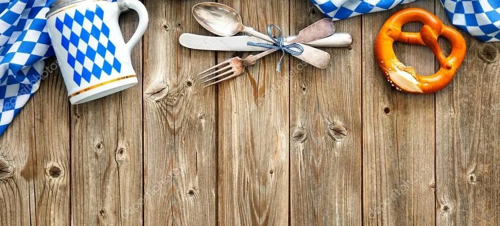 Ein Holztisch mit einem blau-weiß karierten Tuch, einem Bierkrug, einer Gabel, einem Messer, einem Löffel und einer Brezel.
