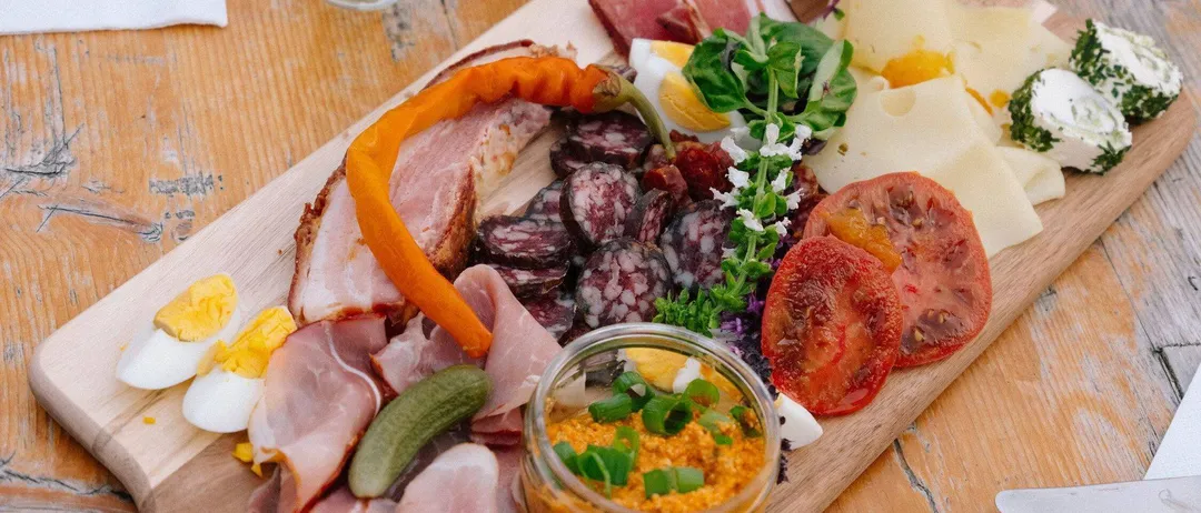 Ein Holzschneidebrett zeigt eine Auswahl an Lebensmitteln, darunter geschnittenes Fleisch, Käse, Tomaten und Gurken.