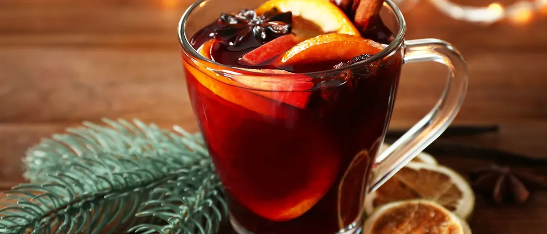 Ein Glas mit rotem Heißgetränk, garniert mit Orangenscheiben, Zimtstangen und Sternanis, auf einem Holztisch mit einem Tannenzweig und einer Vanilleschote.