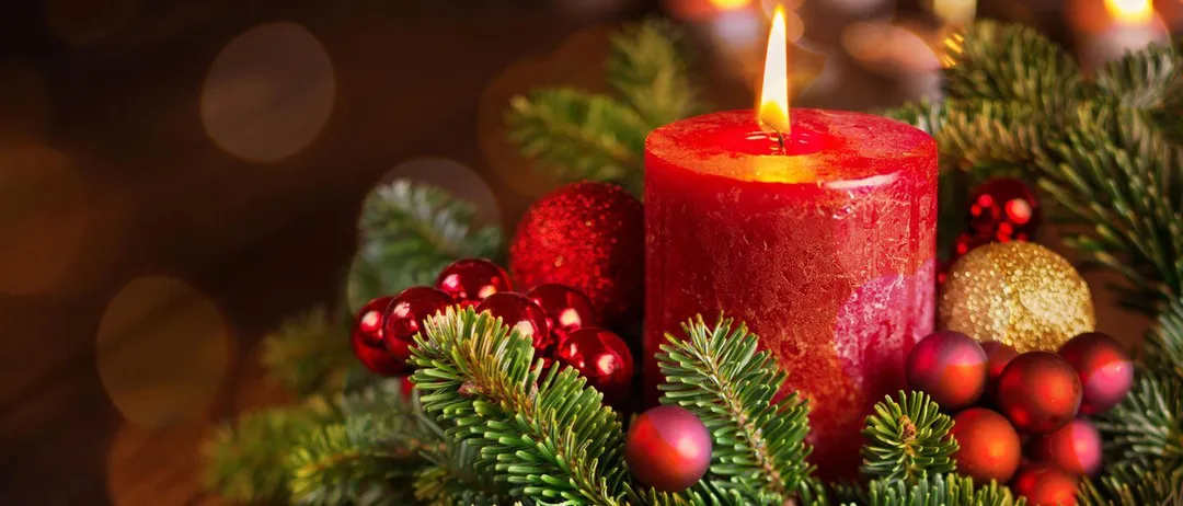 Eine rote Kerze ist unter einem Kranz aus Tannenzweigen und Weihnachtskugeln angezündet.