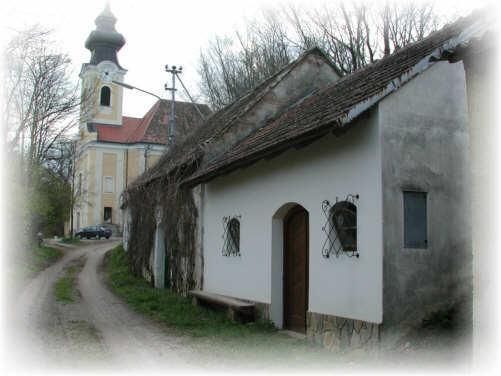 Ein altes Haus mit einem braunen Dach und Efeu an der Wand steht neben einer Straße. Ein Auto ist hinten geparkt. Bäume und ein Kirchturm sind sichtbar.