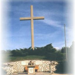 Ein großes Kreuz steht auf einem Hügel, umgeben von einer Steinmauer und Grünpflanzen. Darunter sitzt eine Person an einem kleinen Altar. Der Himmel ist klar und blau.