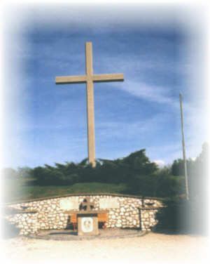 Ein großes Kreuz steht auf einem Hügel, umgeben von einer Steinmauer und Grünpflanzen. Darunter sitzt eine Person an einem kleinen Altar. Der Himmel ist klar und blau.