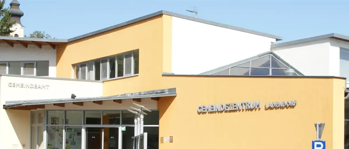 Das Bild zeigt ein gelbes Gebäude mit einem verglasten Eingang. Das Gebäude hat mehrere Fenster und ein Glasdach. Ein Schild mit der Aufschrift 'Gemeindezentrum Ladenburg' ist an der Wand sichtbar.