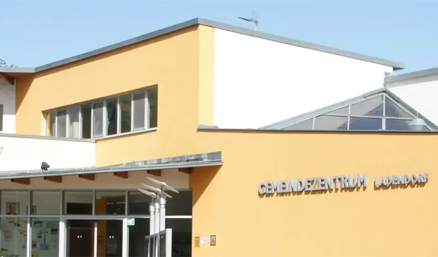 Das Bild zeigt ein gelbes Gebäude mit einem verglasten Eingang. Das Gebäude hat mehrere Fenster und ein Glasdach. Ein Schild mit der Aufschrift 'Gemeindezentrum Ladenburg' ist an der Wand sichtbar.