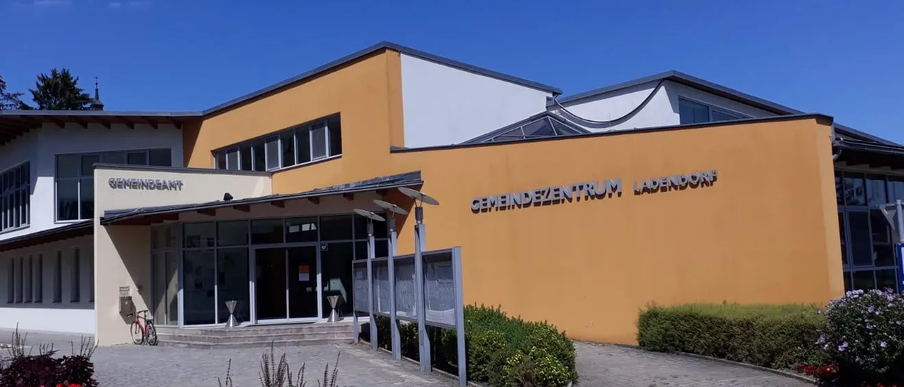 Der Eingang eines modernen Gebäudes mit gelber Fassade und weißen Akzenten. Das Gebäude trägt ein Schild mit der Aufschrift 'Gemeindezentrum Ladendorf'.