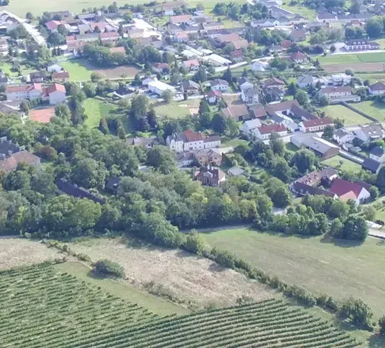 Eine Luftaufnahme einer kleinen Stadt, umgeben von viel Grün, mit zahlreichen Häusern und Gebäuden. Es gibt auch Felder, Bäume und einen Tennisplatz.