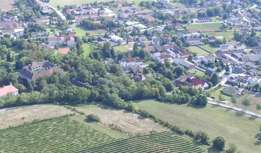 Eine Luftaufnahme einer kleinen Stadt, umgeben von viel Grün, mit zahlreichen Häusern und Gebäuden. Es gibt auch Felder, Bäume und einen Tennisplatz.