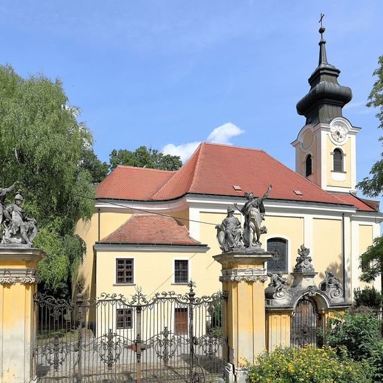 Ein gelbes Gebäude mit einem roten Dach und einem Turm. Das Gebäude hat eine Uhr auf dem Turm. Eine Statue ist auf dem Dach platziert. Das Gebäude hat viele Fenster. Ein Metalltor steht vor dem Gebäude.
