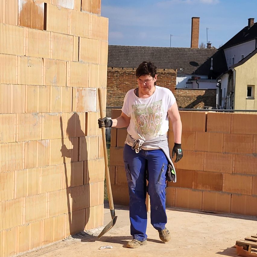Eine Frau mit Brille und Arbeitshandschuhen hält eine Schaufel und steht vor einer Ziegelmauer auf einer Baustelle.