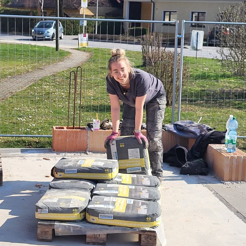 Eine Frau in Arbeitskleidung lächelt, während sie sich auf einen Stapel Zementsäcke auf einem Paletten in einem eingezäunten Außenbereich stützt. Im Hintergrund befindet sich eine Rasenfläche mit einem Haus und ein Auto in der Ferne.