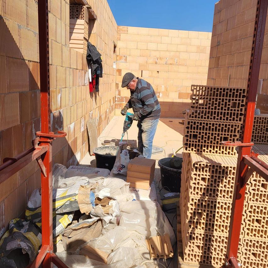Ein Mann bohrt mit einem Bohrer ein Loch in eine Ziegelwand auf einer Baustelle. In der Nähe stehen mehrere Eimer und Ziegel, die auf dem Boden gestapelt sind.