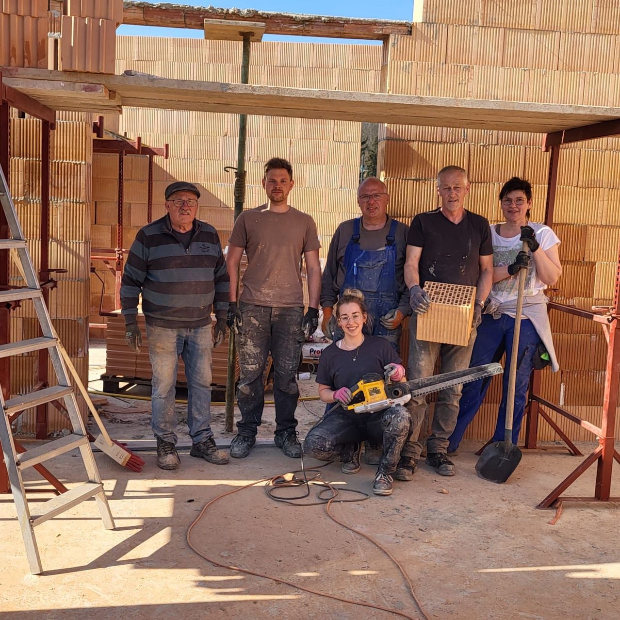 Eine Gruppe von Menschen mit Handschuhen und Brillen posiert für ein Foto auf einer Baustelle. Eine Frau hält eine Kettensäge.