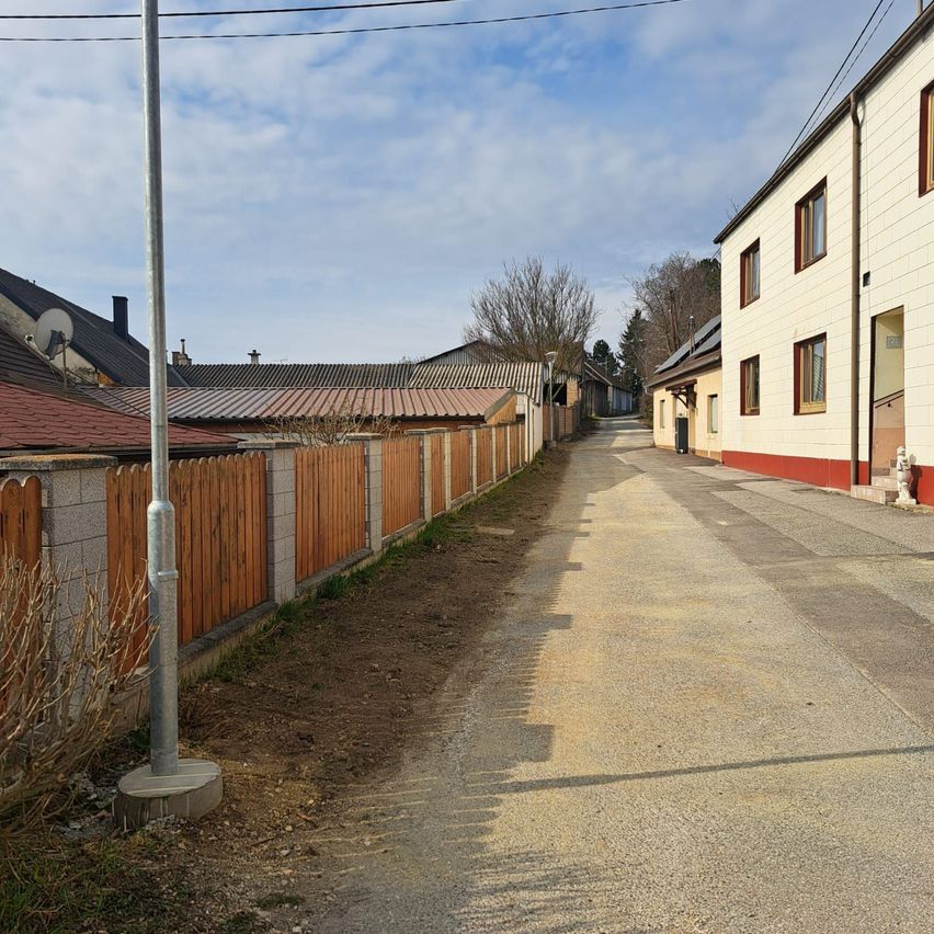 Eine schmale, leere Straße mit Holzzäunen und einem grauen Bürgersteig. Rechts ein weißes Gebäude mit rotem Rand und Treppen. In der Ferne Häuser mit roten Dächern.