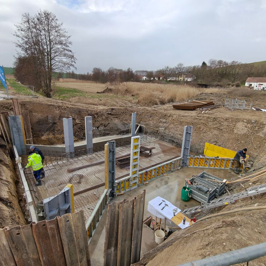 Bauarbeiter arbeiten an einem Gebäudefundament auf einem offenen Feld. Ein Tisch und einige Werkzeuge liegen auf dem Boden. Es gibt Metallbarrieren und Holzzäune rund um das Gelände.