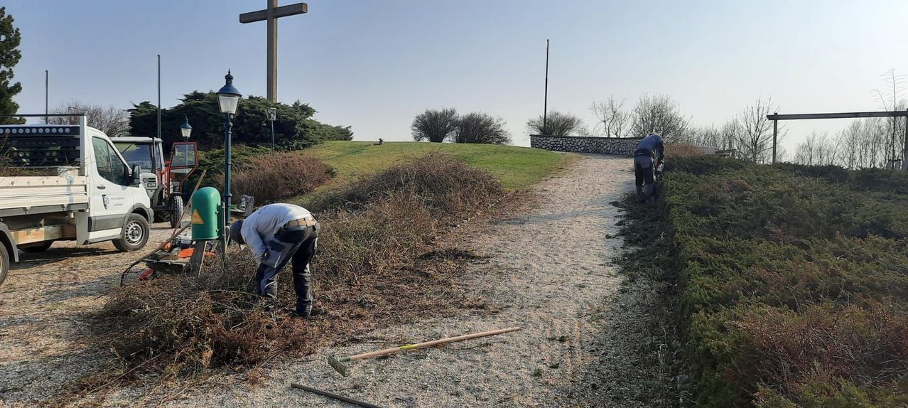 Zwei Personen trimmen Sträucher entlang eines Schotterwegs, wobei ein hoher Kreuz im Hintergrund zu sehen ist.