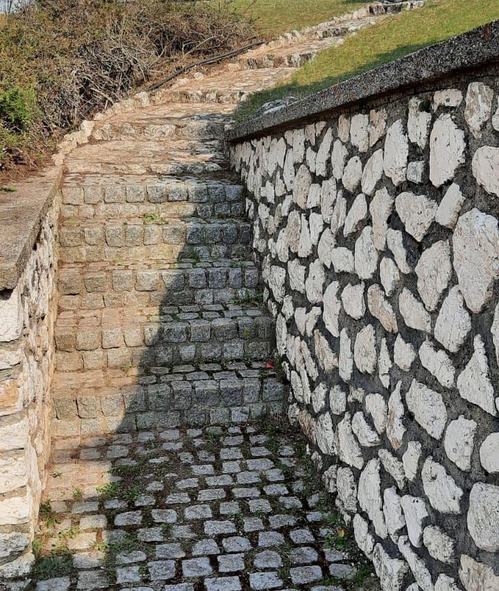 Ein Kopfsteinpflasterweg führt eine Steinmauer hinauf und wirft Schatten auf die Steine. Die Mauer besteht aus großen, unregelmäßigen Steinen und ist von Grün umgeben.
