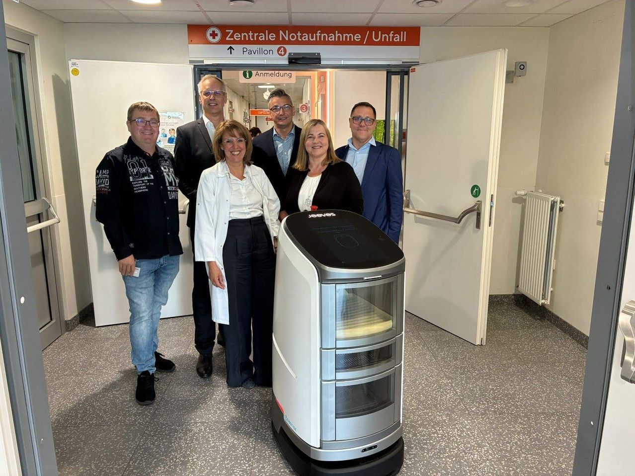 Sechs Personen, darunter drei Frauen, posieren in einem Raum mit einem Roboter und einer offenen Tür. Der Raum hat eine weiße Wand mit einem Schild, das 'Zentrale Notaufnahme/Unfall' steht.
