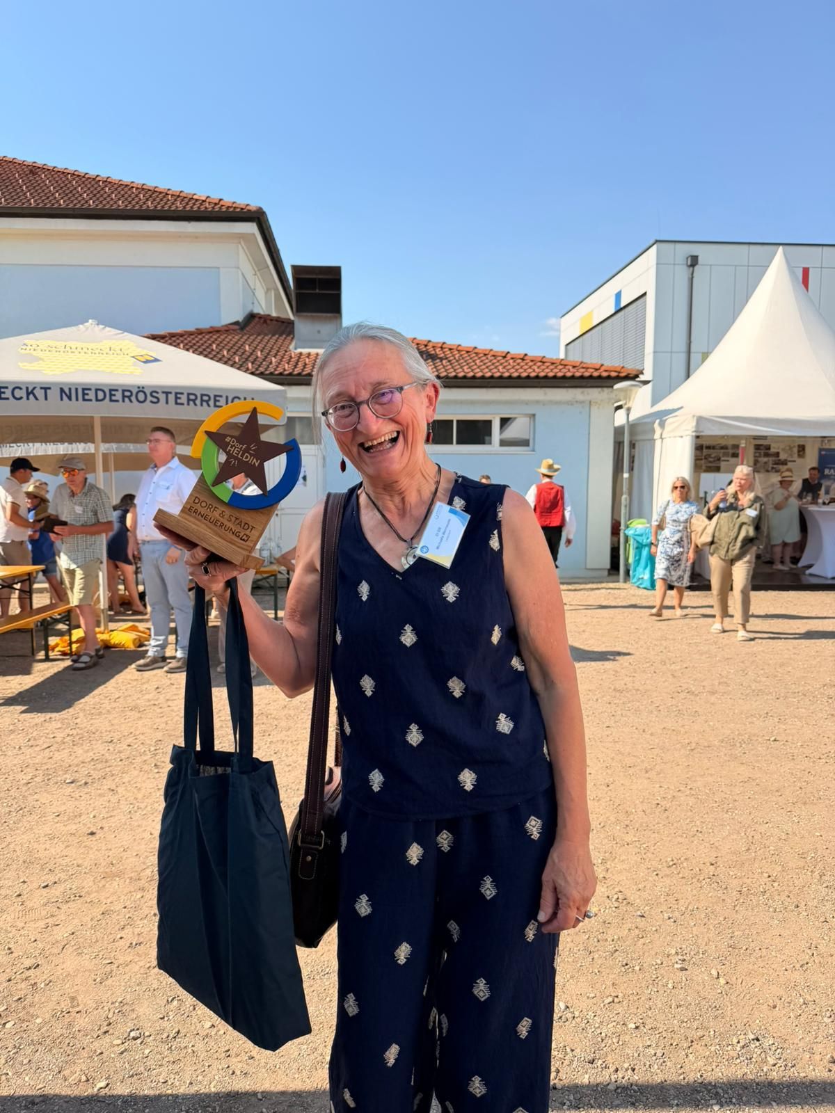 Eine ältere Frau hält einen Pokal und lächelt, steht im Freien. Hinter ihr sind Zelte und Menschen verstreut. Sie trägt ein blaues Kleid mit einem weißen Muster und trägt eine Handtasche.