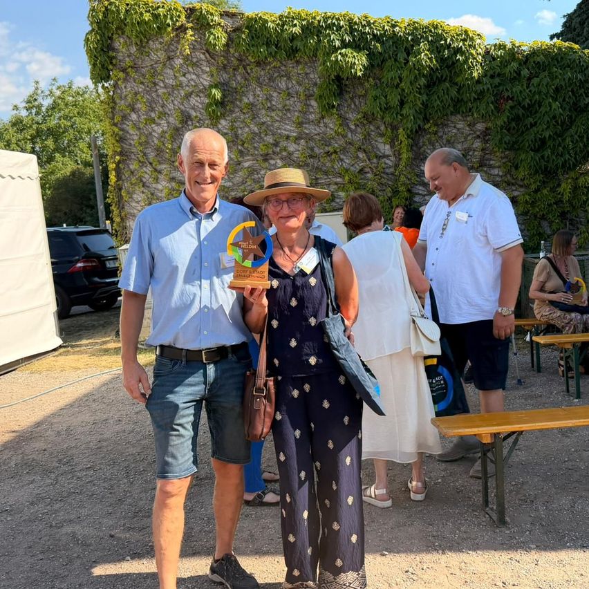 Zwei Erwachsene, ein Mann und eine Frau, stehen mit einem Pokal im Freien. Hinter ihnen sind Bänke und eine mit Efeu bedeckte Steinmauer.