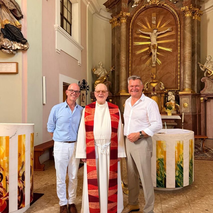 Drei Männer stehen in einer Kirche, einer in geistlicher Kleidung, lächelnd. Hinter ihnen steht ein goldener Altar mit einem Kruzifix und Statuen.