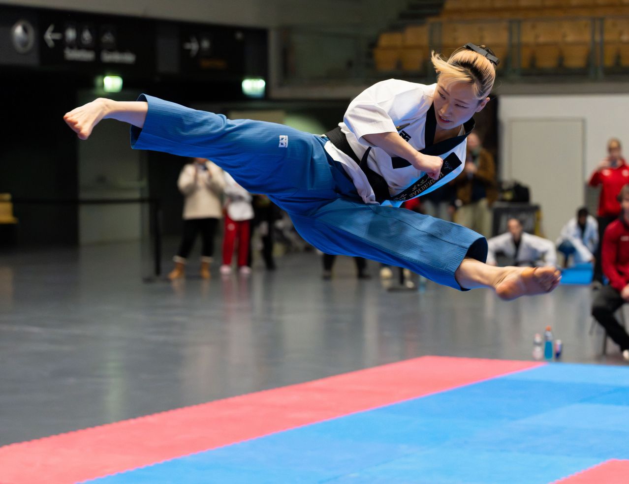 Eine Kampfsportlerin in einem blauen Anzug führt einen hohen Kick in einer Halle aus. Zuschauer beobachten von den Tribünen.