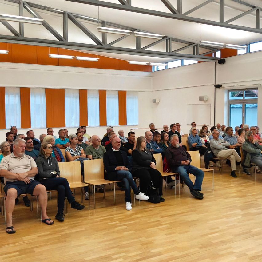 Eine große Gruppe von Menschen sitzt in Stühlen in einem Raum mit Holzböden und großen Fenstern.