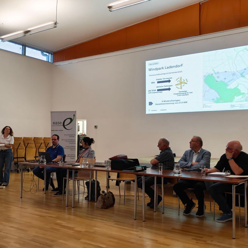 Eine Gruppe von Menschen sitzt an einem Tisch in einem Konferenzraum, mit einem Mann, der vor ihnen steht. An der Wand befindet sich eine Projektionsleinwand, die eine Karte zeigt.