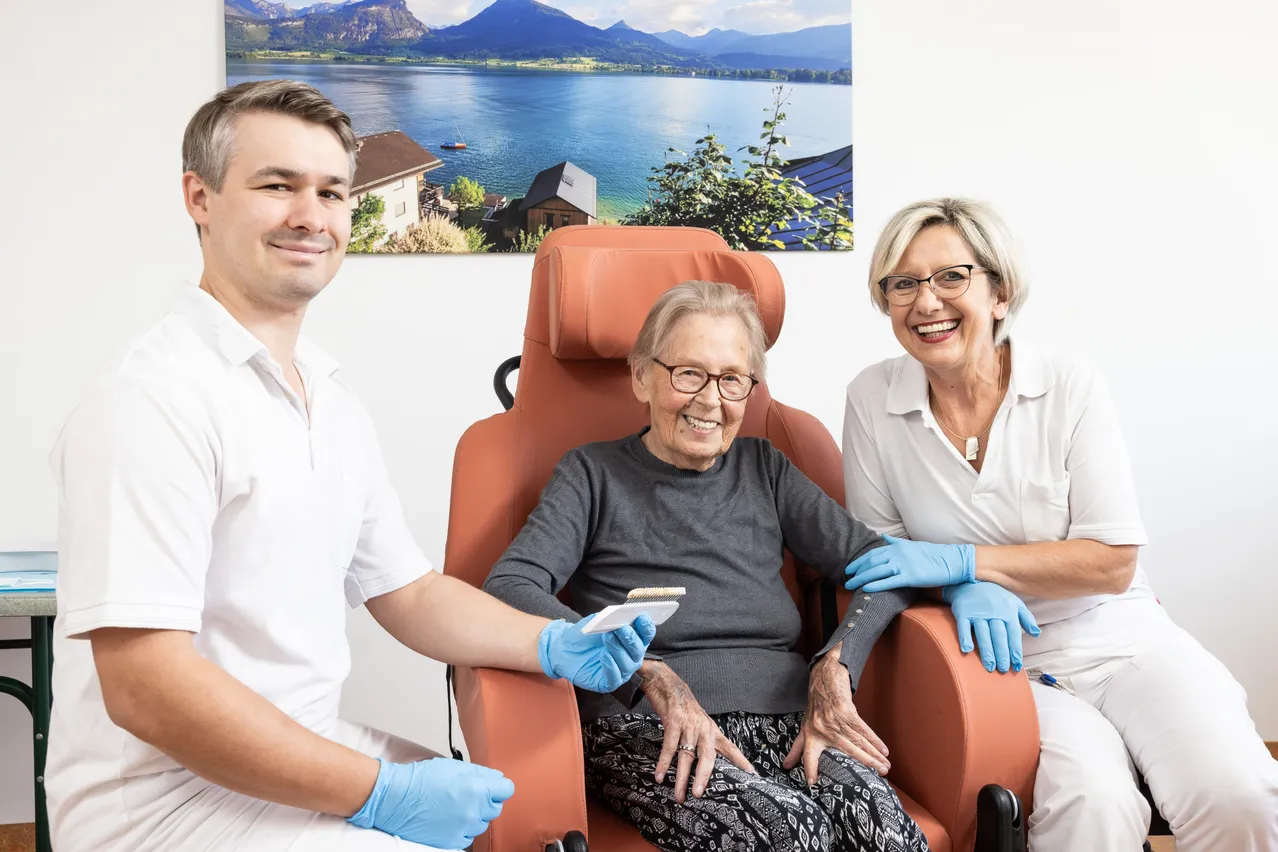 Drei Menschen sitzen in einem Raum. Eine ältere Frau sitzt auf einem Stuhl, während ein Mann und eine Frau neben ihr stehen. Der Mann hält ein kleines Objekt in der Hand.