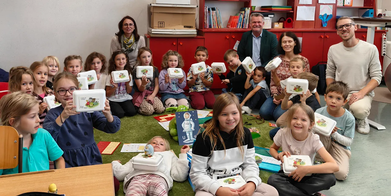 Eine Gruppe von Kindern und Erwachsenen versammelt sich in einem Klassenzimmer. Die Kinder halten Lunchboxen und einige lächeln.