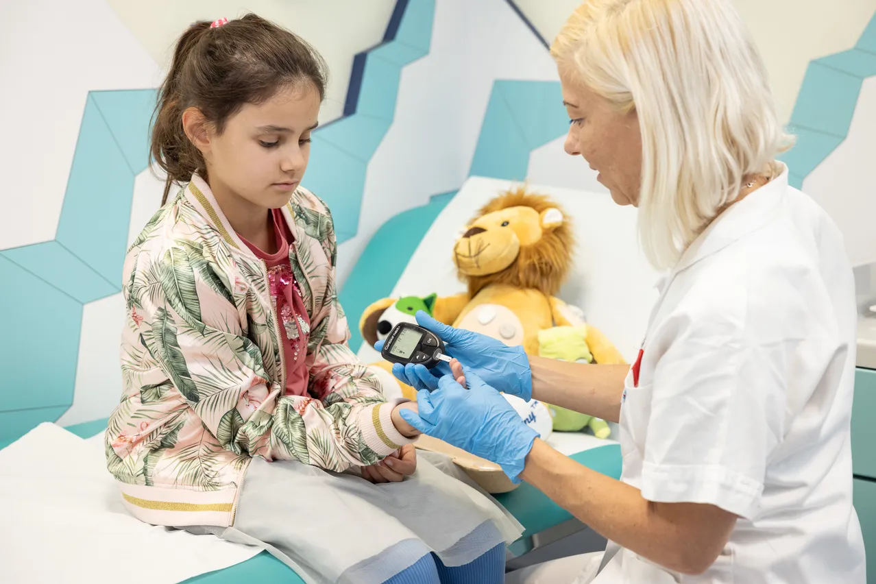 Ein junges Mädchen sitzt auf einem Krankenhausbett, während eine Krankenschwester ihren Blutzuckerspiegel mit einem Glukometer überprüft.