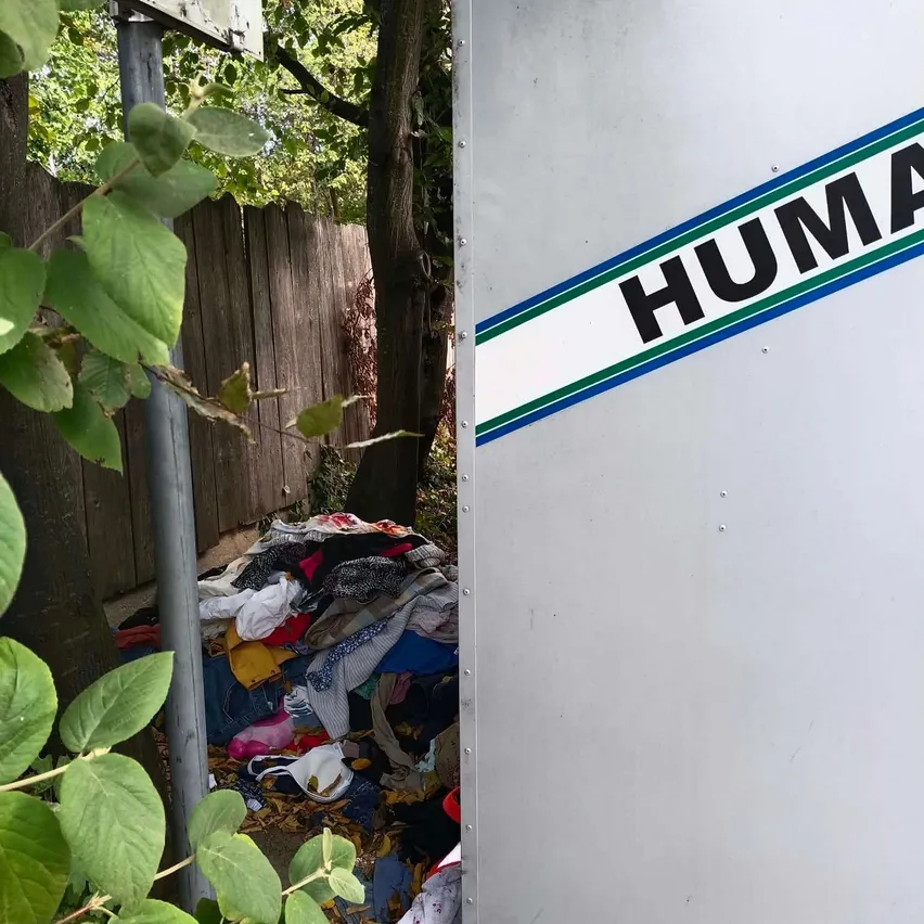 Ein Haufen Kleidung befindet sich neben einem Metallbehälter mit dem Wort 'HUMA' darauf.
