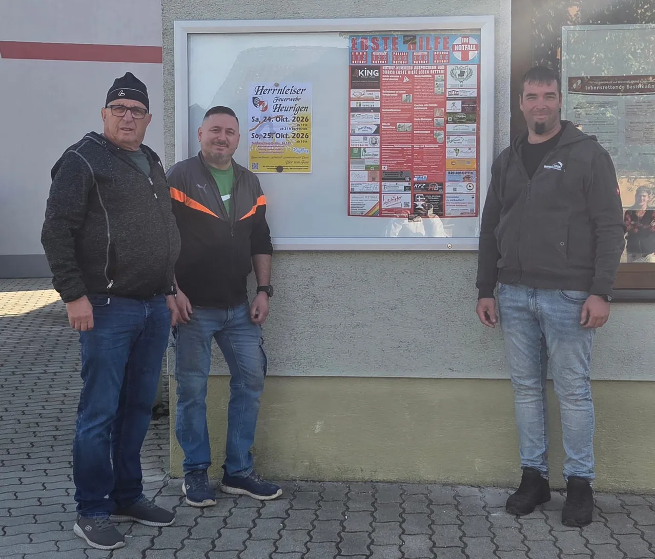 Drei Männer stehen vor einem Gebäude. Der Mann auf der linken Seite trägt eine graue Jacke und Jeans. Der Mann in der Mitte trägt eine schwarze Jacke mit orangefarbenen Streifen und ein grünes Hemd. Der Mann auf der rechten Seite trägt eine schwarze Jacke und Jeans. An der Wand hinter ihnen hängt ein Plakat.