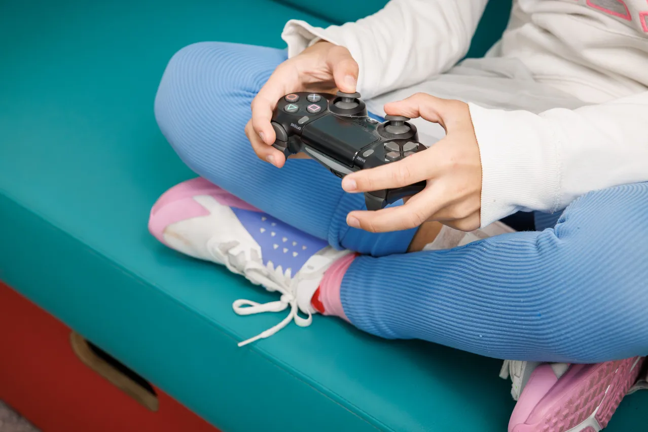 Ein Kind sitzt auf einer blauen Couch und hält einen schwarzen Gamecontroller mit beiden Händen.