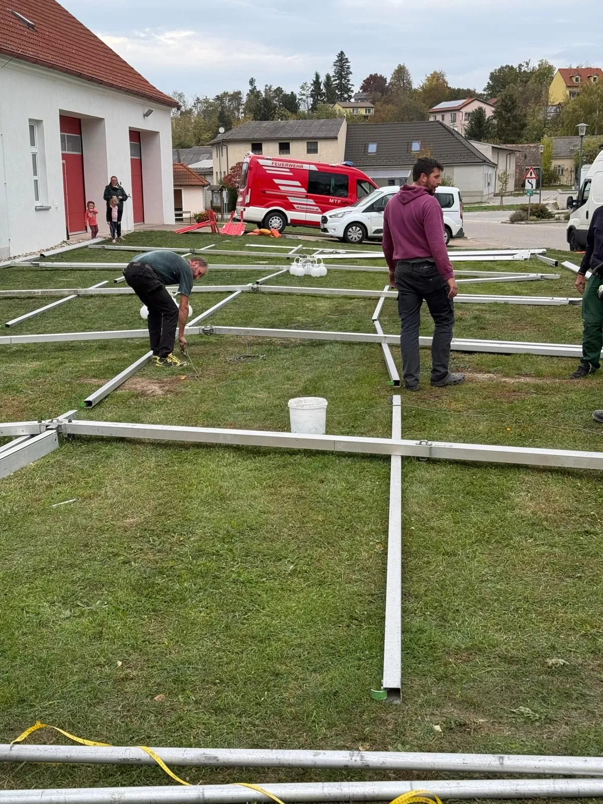 Zwei Männer arbeiten an einer Metallstruktur auf einem Rasen. Ein weißer Eimer steht auf dem Boden. Ein roter Lieferwagen und ein weißes Auto sind geparkt. Ein Gebäude mit roten Türen ist im Hintergrund.