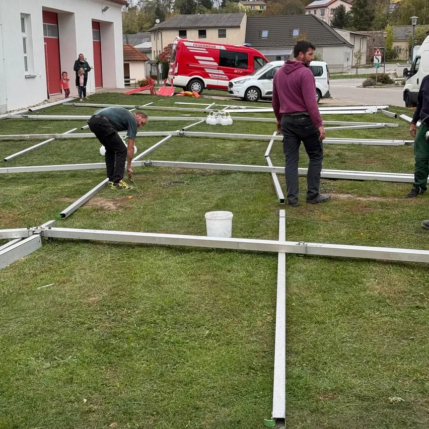 Zwei Männer arbeiten an einer Metallstruktur auf einem Rasen. Ein weißer Eimer steht auf dem Boden. Ein roter Lieferwagen und ein weißes Auto sind geparkt. Ein Gebäude mit roten Türen ist im Hintergrund.