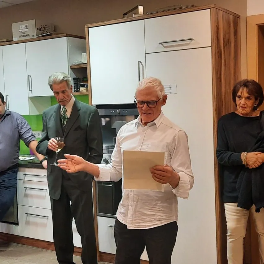 Eine Gruppe von Menschen versammelt sich in einer Küche, wobei ein Mann ein Stück Papier in der Hand hält und zu den anderen spricht.