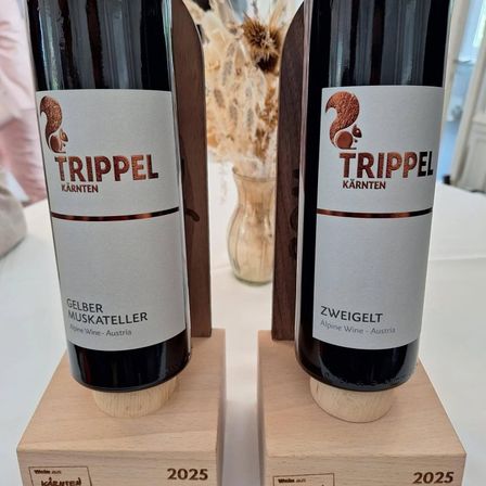 Zwei Flaschen Wein, beschriftet mit 'Trippel', stehen auf Holzsockeln. Die linke Flasche ist 'Gelber Muskateller' aus Österreich, und die rechte ist 'Zweigelt'. Beide stehen auf einer weißen Oberfläche mit einem verschwommenen Hintergrund.