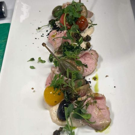 Ein weißer Teller hat ein Gericht mit geschnittenem Fleisch, garniert mit grünem Gemüse, Tomaten und schwarzen Oliven, mit Sauce beträufelt und schwarzem Pfeffer obenauf.