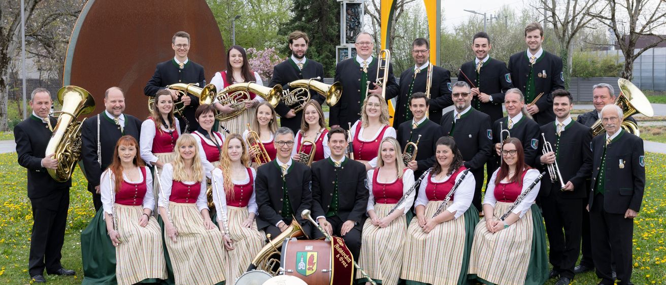 Eine Gruppe von Musikern, gekleidet in traditioneller Tracht, posiert für ein Foto im Freien. Sie halten verschiedene Instrumente. Hinter ihnen steht eine große runde Skulptur und eine gelbe bogenförmige Struktur.