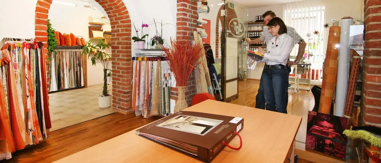 Ein Paar untersucht ein Buch auf einem Holztisch in einem Geschäft, mit dekorativen Pflanzen und einer Ziegelwand im Hintergrund.