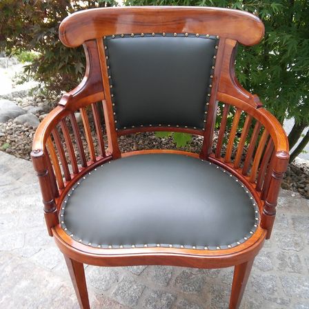 Ein hölzerner Sessel mit schwarzem Ledersitz und -rückenlehne, verziert mit goldenen Nieten, auf einer Steinfläche mit Steinen und Pflanzen im Hintergrund.