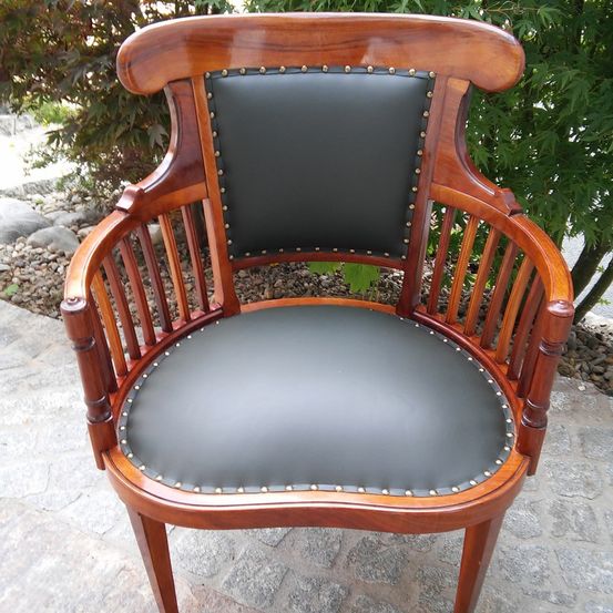 Ein hölzerner Sessel mit schwarzem Ledersitz und -rückenlehne, verziert mit goldenen Nieten, auf einer Steinfläche mit Steinen und Pflanzen im Hintergrund.