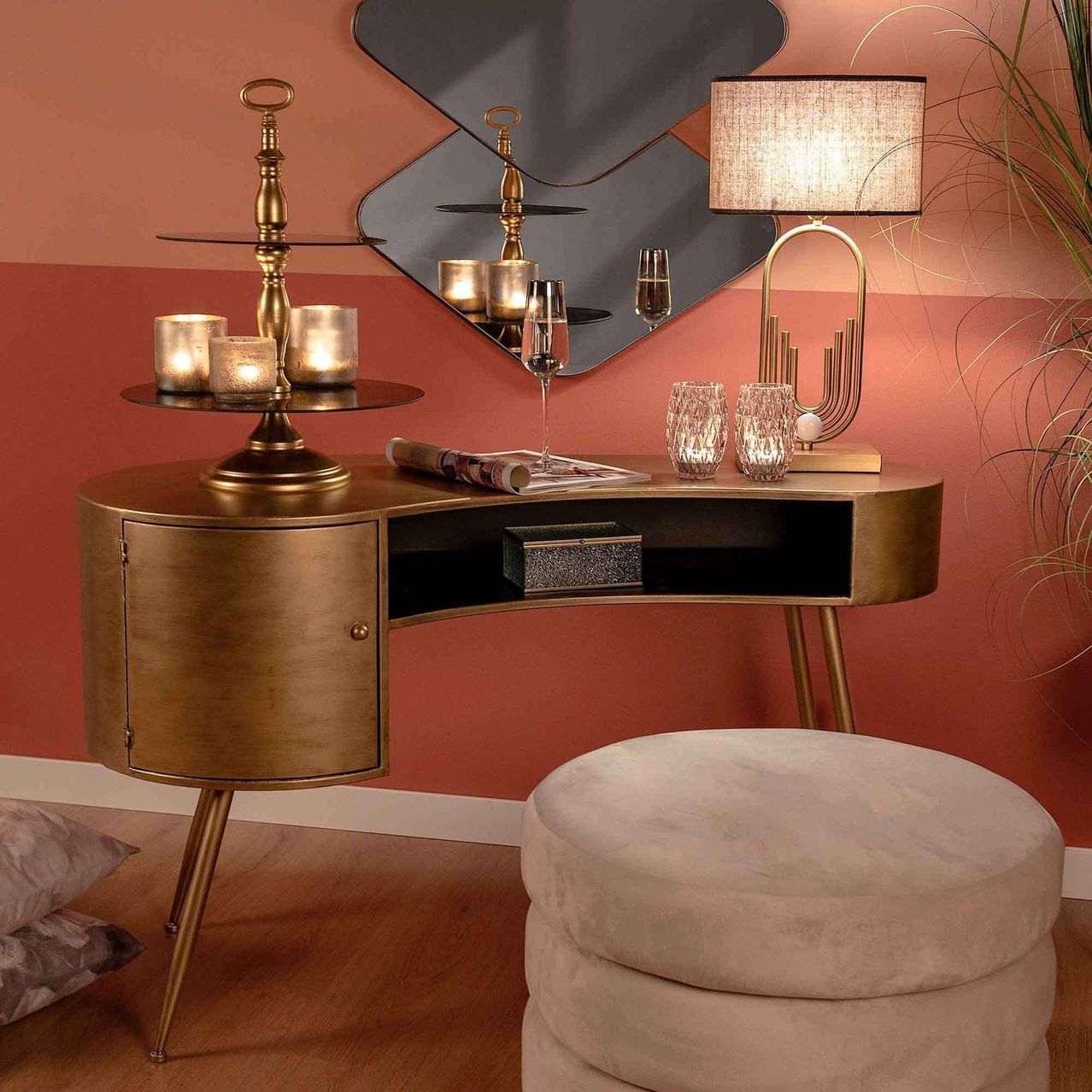 A stylish vanity desk with brass accents stands in a room, featuring a lamp, a stack of candles, and a magazine. A mirror reflects the room, and a cushioned stool sits nearby.