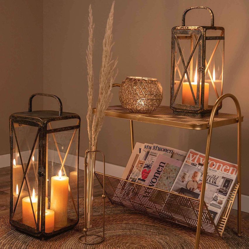 A cozy corner with a lit lantern, candles, and magazines on a tray. A tall plant is in a vase on the floor.