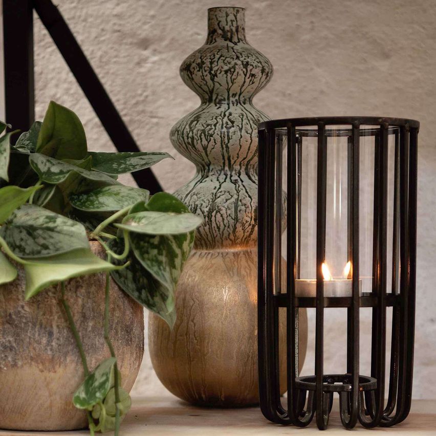 Three decorative objects on a wooden surface, including a plant in a pot, a tall vase, and a candle holder with a lit candle.