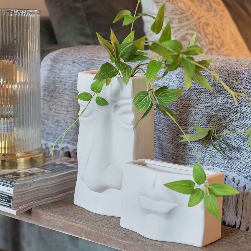 Two white ceramic vases with human face designs, holding green plants, sit on a wooden shelf next to a glass lamp.